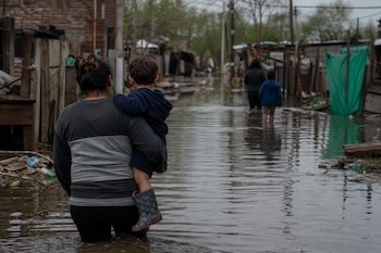 Una mujer carga a un niño mientras avanza por un camino inundado flanqueado por viviendas precarias.