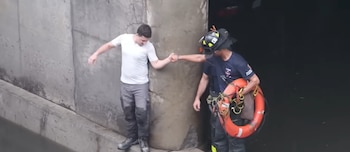 Bombero con casco y salvavidas naranja ayuda a un hombre a subir de una zona inundada con pared de cemento y agua oscura