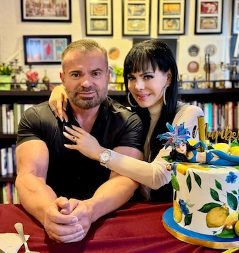 Maribel Guardia y un hombre posan sonrientes detrás de un pastel blanco decorado con limones, hojas, flores azules y una figura de mujer con tocado de plumas