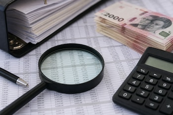 Planilla con números examinada con una lupa junto a un bolígrafo, una calculadora negra, una carpeta con hojas y fajos de billetes de 2000 pesos.