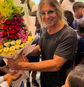 Un hombre con anteojos y cabello largo sostiene un gran ramo de flores coloridas rodeado por personas