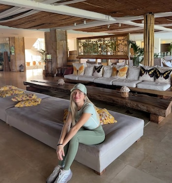 Laurita Fernández sentada sonriente en un banco de tela gris en el lobby de un hotel, vistiendo un conjunto deportivo verde y gorra, con muebles rústicos detrás