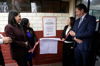 Cuatro personas con vestimenta formal frente a una placa conmemorativa en una pared de ladrillo. Sujetan una cinta blanca y roja
