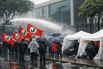 Manifestantes con impermeables y paraguas bajo un chorro de agua de cisterna. Banderas rojas, carpas blancas y charcos reflejan el edificio al fondo.