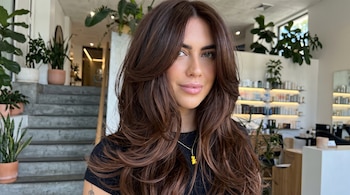 Mujer de cabello largo castaño peinado en capas en un salón de belleza con plantas y estantes con productos.