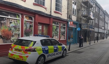 Coche de policía blanco y azul estacionado en una calle empedrada con tiendas al fondo. Un oficial de policía está de pie cerca de un edificio con andamios