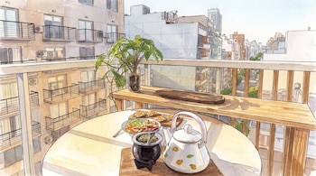 Ilustración al estilo acuarela de una mesa de balcón con un recipiente de mate, una pava, tostadas y fruta fresca con edificios al fondo.