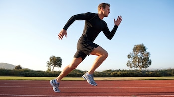 Hombre caucásico musculoso corriendo en pista de atletismo. Viste camiseta térmica negra de manga larga, pantalones cortos negros y zapatillas deportivas grises.
