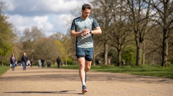 Un hombre corre por un sendero de tierra en un parque, con camiseta gris y shorts azules, mientras mira su reloj en la muñeca.