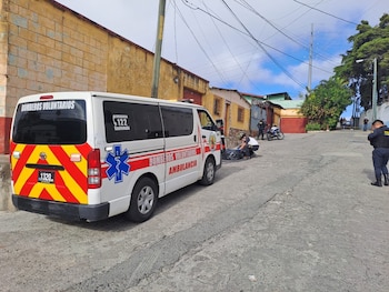 Ambulancia tipo van blanca, personal de emergencia agachado, objeto negro en el suelo, policías, motocicleta, calle y edificios