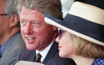 Primer plano de Bill Clinton con Hillary Clinton a su derecha. Él viste traje oscuro, camisa blanca y corbata multicolor. Ella usa gafas de sol y un sombrero de paja