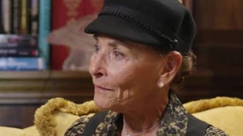 Judy Sheindlin de perfil con una gorra negra, un arete de perla y una prenda con estampado, ante una estantería
