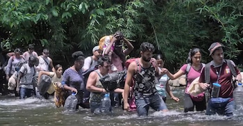 Grupo de personas camina en un río. Llevan mochilas y botellas de agua. Hay árboles y vegetación densa en el fondo. Un adulto carga a un niño