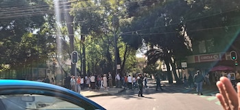 Vista de una calle con personas reunidas en la acera, árboles frondosos, edificios al fondo con un letrero de 'Circle K' y un semáforo en verde
