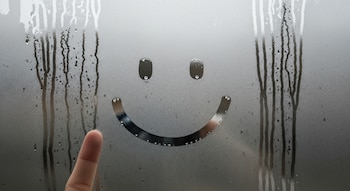 Vidrio empañado con condensación y gotas de agua. En el centro, una cara sonriente dibujada. Un dedo humano aparece en la parte inferior izquierda.