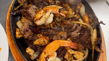 Primer plano de una sartén de hierro fundido humeante con trozos de carne asada, camarones anaranjados y rodajas de cebolla blanca