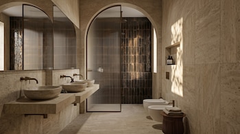 Un baño de estilo moderno con paredes de travertino, dos lavabos de piedra sobre encimera, grifos de bronce, una ducha y un inodoro con bidé.