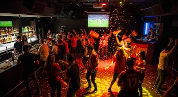 Personas celebrando en una discoteca con banderas de España, una pantalla de televisión, una barra con bebidas y confeti en el suelo.