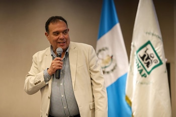 Hombre con camisa a cuadros y saco beige sostiene un micrófono. Detrás, una bandera de Guatemala y una bandera blanca con el logo de una organización
