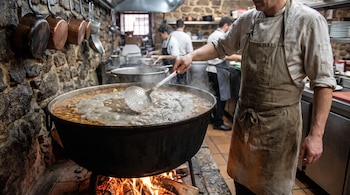 Un cocinero con delantal usa una espumadera en una olla grande sobre brasas, con sartenes en la pared y personal al fondo.