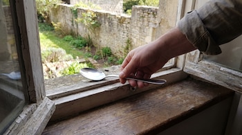 Primer plano de una persona colocando una cuchara de acero inoxidable en el marco de una ventana, con el mango hacia adentro y el cuenco hacia afuera.