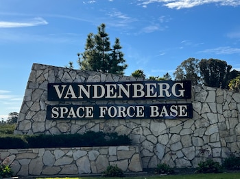 Un cartel de piedra con la inscripción Vandenberg Space Force Base en letras blancas sobre fondo negro, rodeado de vegetación bajo un cielo azul