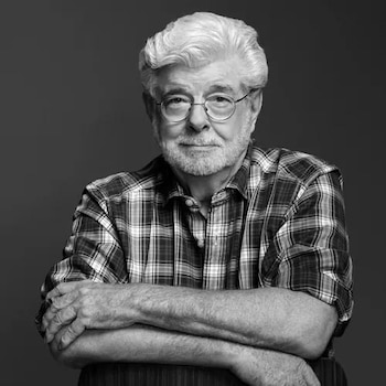 Retrato en blanco y negro de George Lucas, un hombre blanco con cabello y barba blancos, gafas redondas, camisa a cuadros, con los brazos cruzados