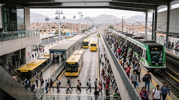 Estación de transporte intermodal con buses amarillos, un tren urbano, pasajeros en los andenes y cabinas de teleférico al fondo