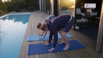 Un hombre y una mujer practican yoga en colchonetas azules sobre un deck de madera, junto a una piscina y un edificio con puertas de vidrio