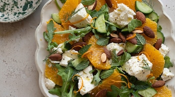 Primer plano de una ensalada fresca con rodajas de naranja, trozos de queso blanco, pepino, almendras enteras y hojas verdes de rúcula y menta, sobre un plato blanco.