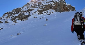 Un montañista de espaldas con chaqueta roja y mochila camina sobre la nieve en dirección a una cumbre rocosa
