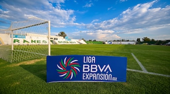 Campo de fútbol con césped verde, una portería blanca, un cartel azul con el logo Liga BBVA Expansión y gradas blancas con la palabra Durango, cielo azul.