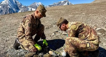 Dos soldados con uniforme de camuflaje y gorra revisan con una cinta métrica un proyectil sobre el suelo rocoso de una montaña
