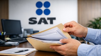 Manos de una persona sostienen una carpeta abierta con papeles. En el fondo, el logo SAT, un escritorio con computadora y bolígrafo.