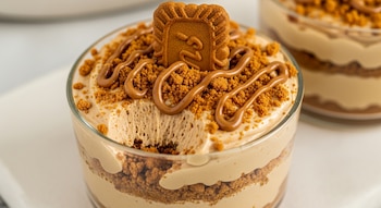 Primer plano de un postre Lotus sin horno en un vaso de cristal, con capas de crema, galletas trituradas, salsa Biscoff y una galleta Lotus entera encima.