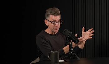 Un hombre de mediana edad con gafas y camisa oscura habla en un micrófono negro, gesticulando con ambas manos. Una taza oscura está sobre la mesa negra