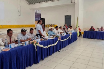 Un grupo de hombres y mujeres sentados en dos mesas largas con manteles azules, botellas de agua y papeles. Un hombre de pie habla por micrófono. Hay un cartel y una bandera en el fondo