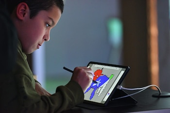 Un niño de perfil dibuja con un lápiz digital en una tableta horizontal sobre una mesa, mostrando un diseño de formas coloreadas en rojo, azul y verde