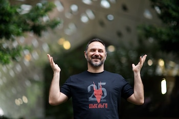 Hombre con barba y camiseta negra con un diseño rojo y gris que levanta ambas manos en un espacio con fondo desenfocado