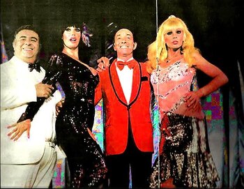 Cuatro personas posan: hombre en traje blanco, mujer en vestido negro brillante, hombre en saco rojo y mujer rubia en top rosa y falda brillante