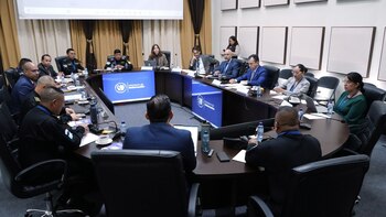 Un grupo diverso de personas, incluyendo oficiales y civiles, sentadas en una gran mesa ovalada durante una reunión. Se observan pantallas con el logo del Ministerio de Gobernación