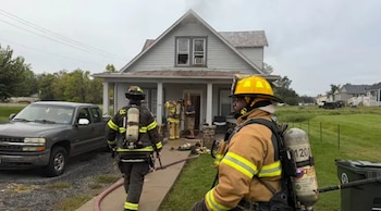 Bomberos con cascos y equipos de respiración caminan hacia una casa de madera junto a una camioneta gris