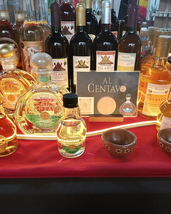 Múltiples botellas de vino y mezcal de distintas variedades, una botella pequeña de mezcal blanco y copas de cerámica, sobre un mantel rojo