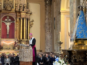 El arzobispo Jorge García Cuerva, con sotana negra y fucsia, habla desde un púlpito de metal dorado en una iglesia, con fieles y estatuas religiosas de fondo