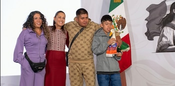 Una mujer y Claudia Sheinbaum junto a dos jóvenes, uno de ellos sostiene un pato de juguete y un pañuelo de FIFA, con una bandera de México detrás