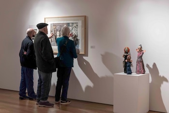 Tres personas de espaldas, dos hombres y una mujer, observan una pintura enmarcada en una pared blanca, con tres figuras de estilo marioneta sobre un pedestal a la derecha