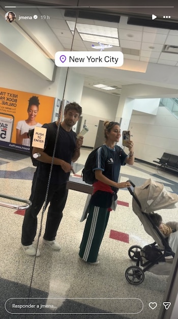 Una mujer y un hombre se reflejan en un vidrio con un cochecito de bebé en un aeropuerto. El hombre sostiene un boleto y la mujer se toma una selfie