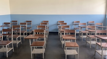 Un aula vacía con filas de pupitres de madera y metal blanco. Las paredes son de color azul claro en la parte inferior y crema en la superior. El suelo es oscuro.