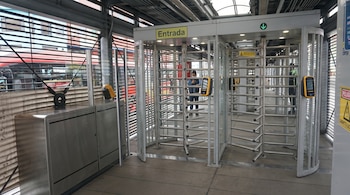 Torniquetes de piso a techo en estación de TransMilenio. Un pasajero con mascarilla usa el validador. Un bus rojo es visible en el exterior.