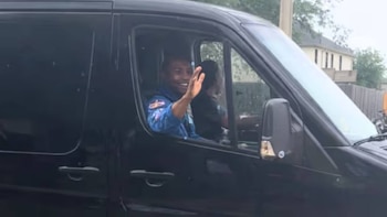 Hombre de tez oscura en uniforme azul con parches en el brazo saluda desde la ventanilla abierta de una van negra. Se ve parcialmente al conductor